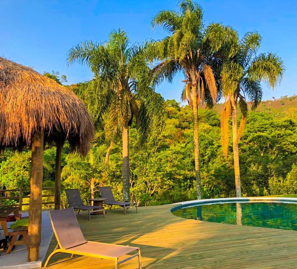 Deck de madeira localizado á beira da piscina com um quiosque de sapê localizado ao lado esquedo.