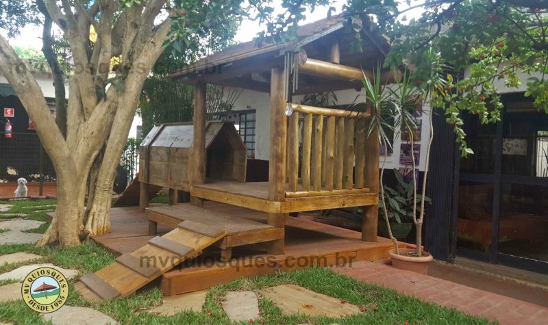 Uma casinha de cachorro com uma varanda e uma rampa para o animal subir, com uma árvore ao lado.